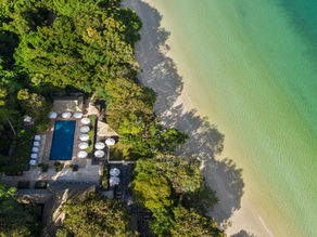 Aerial view of a serene beachfront with turquoise water, lush green trees, and a pool surrounded by white umbrellas and lounge chairs.