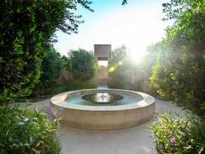 Circular stone fountain with water feature surrounded by lush greenery and bushes. Sunlight filters through trees, creating a serene mood.