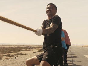 Man in black shirt and gloves pulls rope on road with others. Sunny day, desert landscape. Determined expression, team effort.