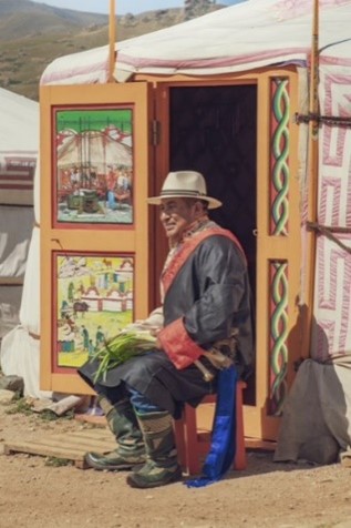 Smiling man in traditional hat and robe sits outside yurt