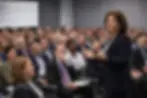 Female executive leader in her 50s speaking confidently to a large, diverse audience of staff at a corporate retreat, engaging attendees who are listening attentively and taking notes.