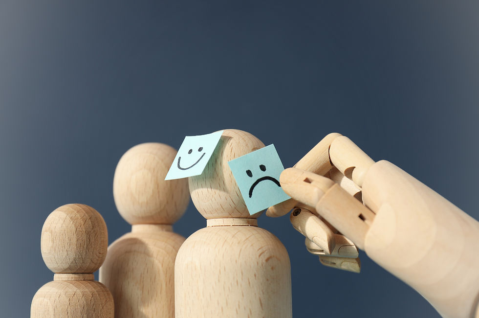 A wooden mannequin hand replaces a sad face sticky note with a happy face on the head of a wooden figure, standing among other faceless wooden figures, symbolizing emotional transformation or support for mental health.