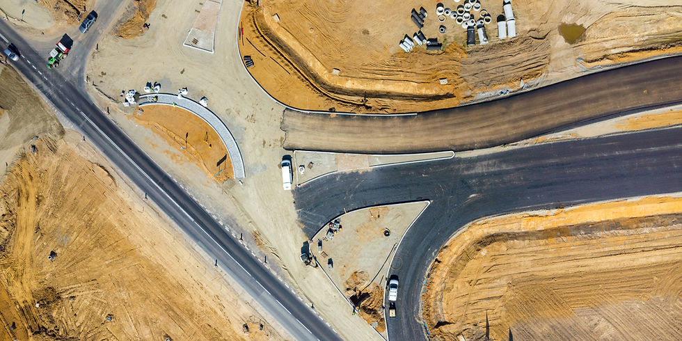 Aerial view of road construction site with vehicles, equipment, brown dirt, and asphalt. Workers in safety gear visible on the curved road.