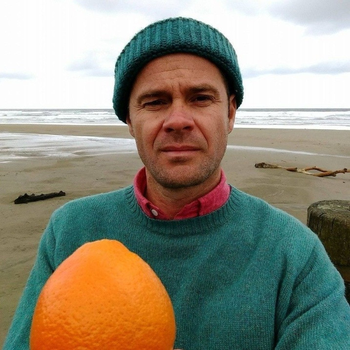 Featured author Roger Peet posing on beach with an orange.