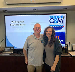 A man and a woman stand in front of a presentation screen. The screen displays the words "Working with DeafBlind Riders" as well as the logo of a company called "Shore O&M, Orientation & Mobility." The man is wearing a light blue polo shirt with a small logo on the left side of his chest, khaki pants and a smile. The woman is wearing a grey long-sleeved shirt and a dark green cardigan. She is also smiling. The presentation screen is on top of a brown wooden cabinet. There is a laptop, a remote and a black water bottle on the cabinet.  The presentation screen is in a room with beige walls.  The image is likely a photo taken during a training session for working with deafblind riders.