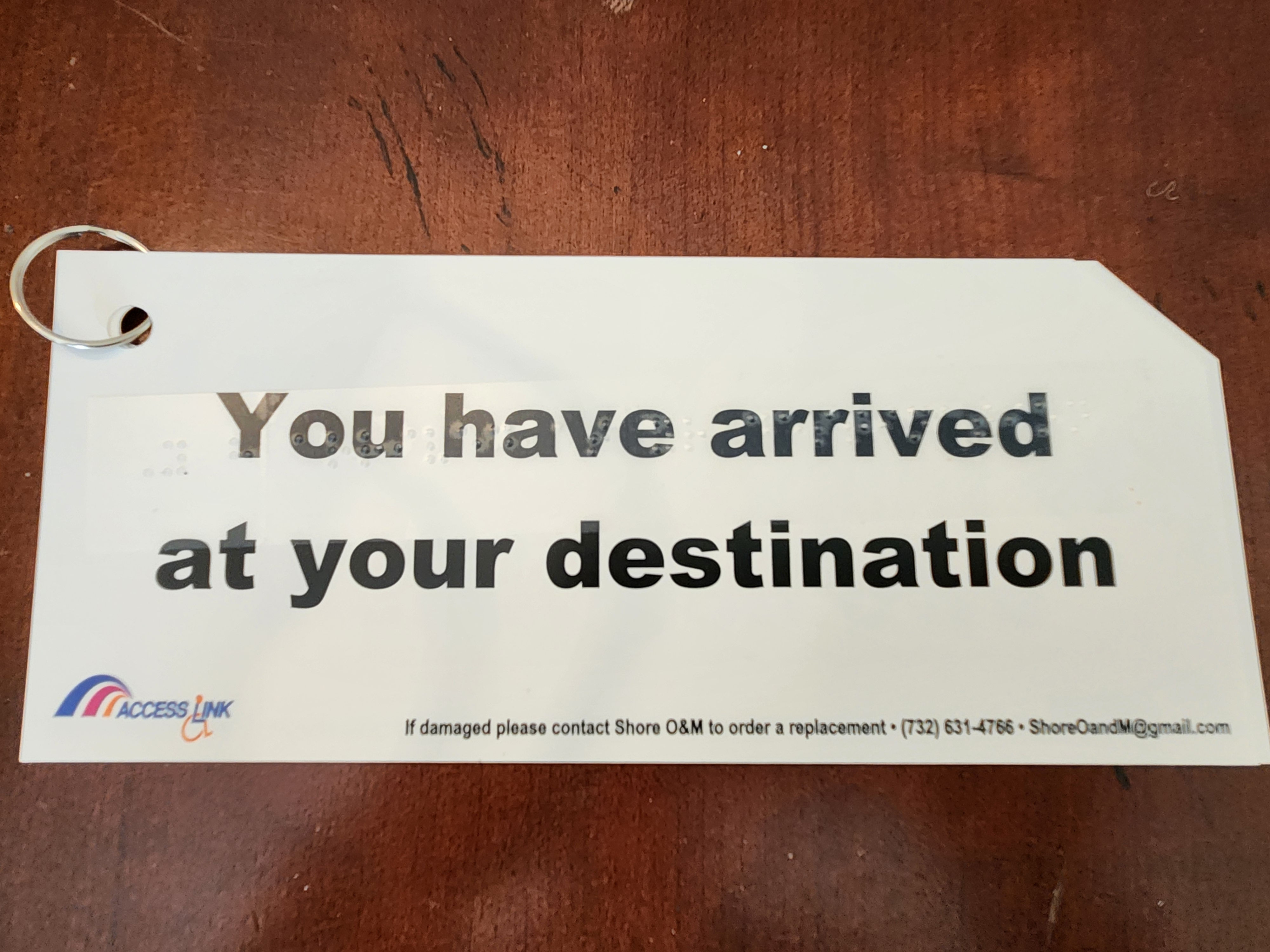 a rectangular white card with one clipped corner. The card is attached to a metal ring in the top left corner. It is placed on a wooden surface, which shows a textured grain pattern. The card features bold black text reading, "You have arrived at your destination." Below the printed text, there is a strip of clear tape embossed with Braille, paralleling the printed message.