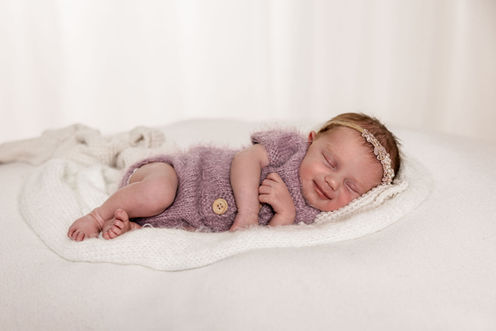 Schlafendes und lächelndes Neugeborenes in einem lila Strickoutfit bei einem professionellen Newbornshooting