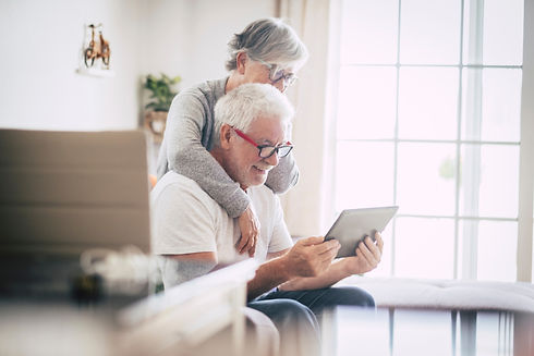 stock photo of older couple. The husband is sitting down reading a tablet whiel the wife is standing behidn him, with her arms around his shoulders