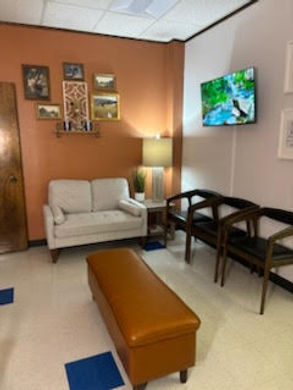 a waiting room or reception area which includes a comfortable two-seater sofa, a long brown ottoman, and three dark wooden chairs