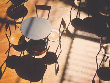 Shadows cast by sunlight on a wooden floor, highlighting a round table surrounded by orange chairs, creating a warm, inviting ambiance.