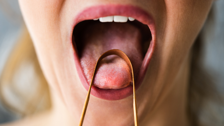 A close-up of a person scraping their tongue with a metal tongue scraper, mouth open wide to show the tongue being cleaned.