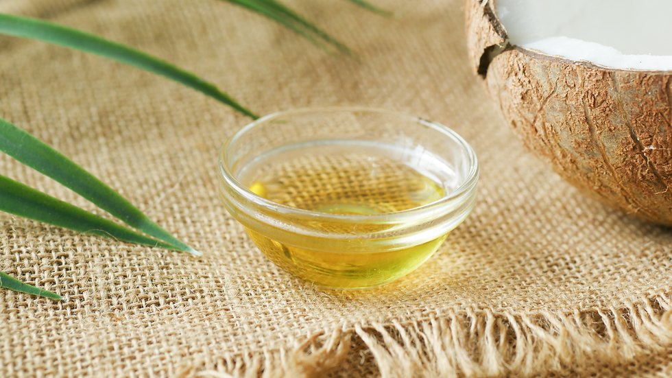 Small glass bowl filled with light golden oil placed on burlap fabric, with part of a cracked coconut and green leaves nearby.