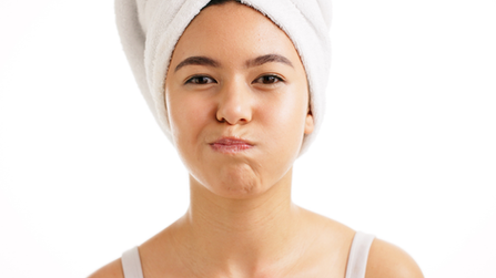 A young woman with a towel wrapped around her head puffs out her cheeks as if swishing liquid in her mouth, standing against a white background.