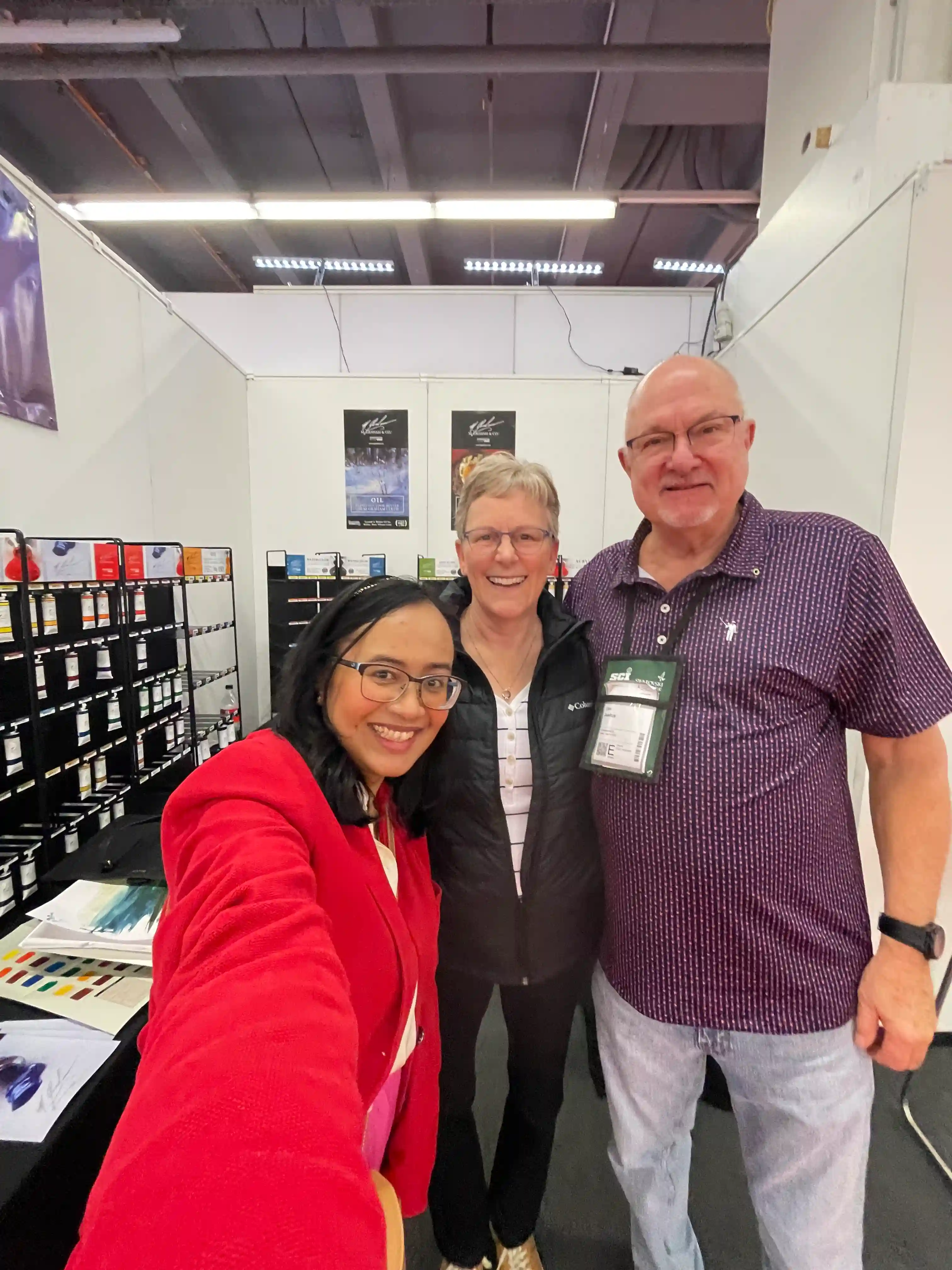 Artist Fizah at the M. Graham booth during Creativeworld Messe, standing with Judy Rebitzke and Dan Justus. The photo captures a moment of discussion about M. Graham’s artist-grade watercolor paints, known for their rich pigments and smooth application. A lively trade fair setting with art supplies displayed in the background.
