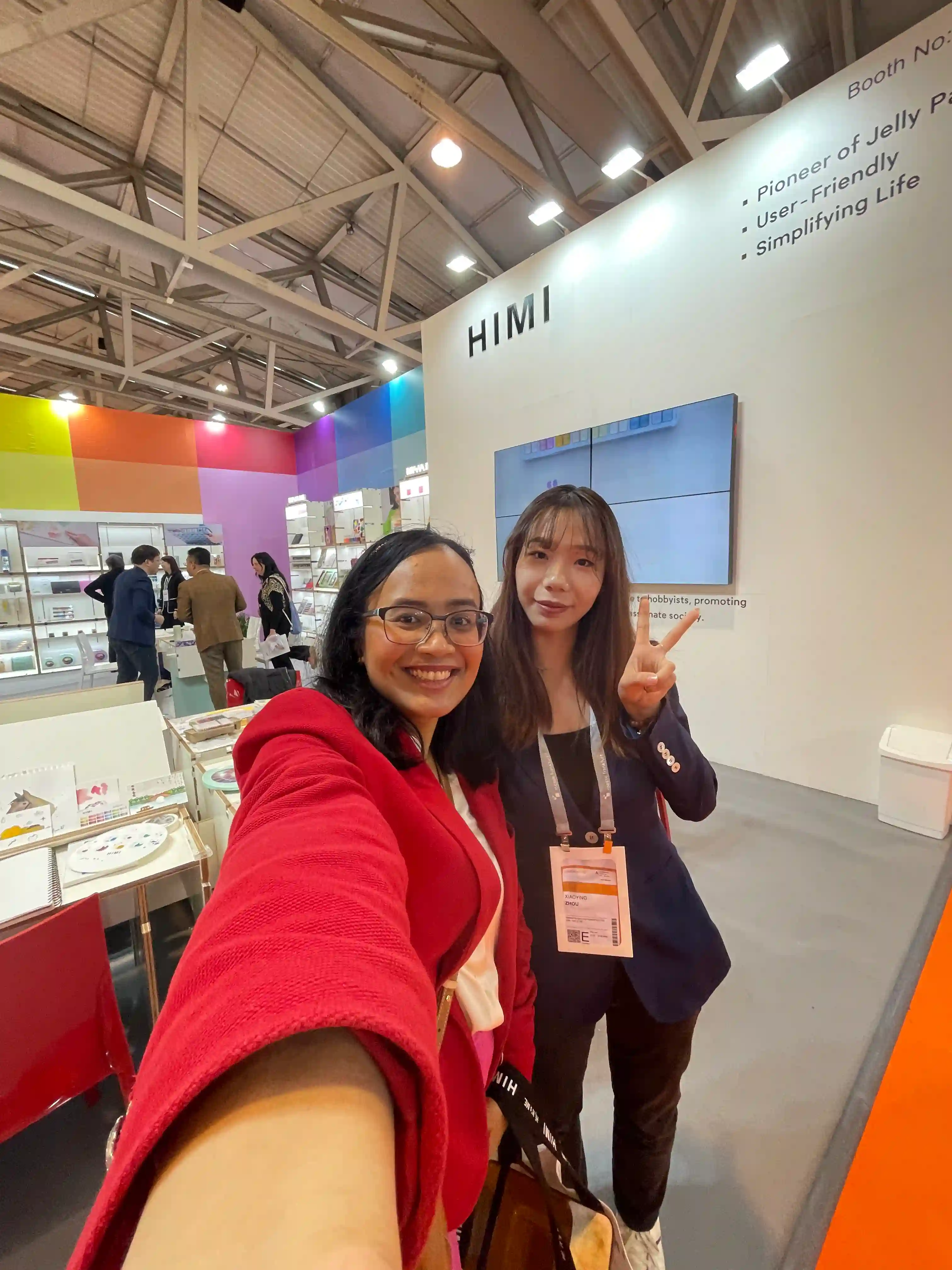Artist Fizah visiting the HIMI booth at Creativeworld Messe, standing with Michelle Zhou. The photo captures a moment of discussion about HIMI’s signature gouache paints, known for their vibrant pigments and smooth, creamy consistency. Behind them, colorful displays of art supplies add to the creative atmosphere of the trade fair.