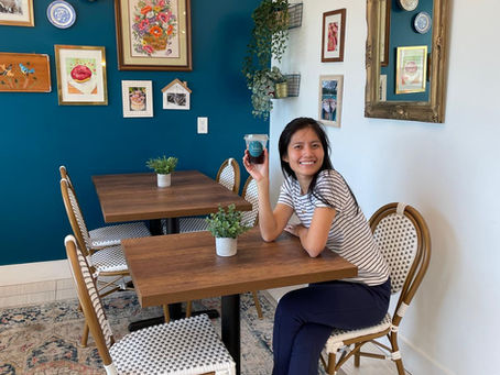 Smiling bakery owner with a coffee, seated at a café table with green plants, against a blue wall adorned with framed art and mirror.