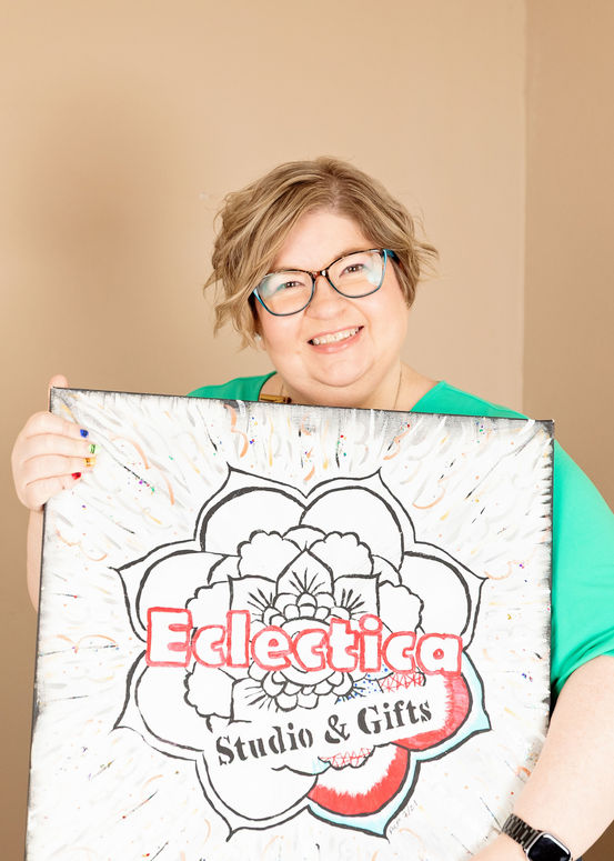 Smiling woman wearing glasses holds a hand-drawn Eclectica Studio & Gifts logo canvas with floral design.