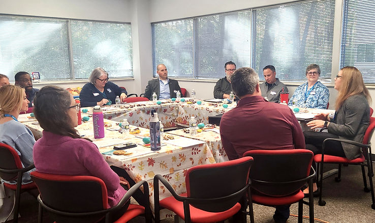 Meeting in a room with 12 people seated around tables with autumn-themed tablecloths. Lively discussion, water bottles, and snacks visible.