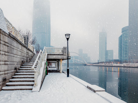 Snowy urban scene with snowy steps and lamppost along a river. Skyscrapers in the misty background. Quiet, wintry atmosphere.