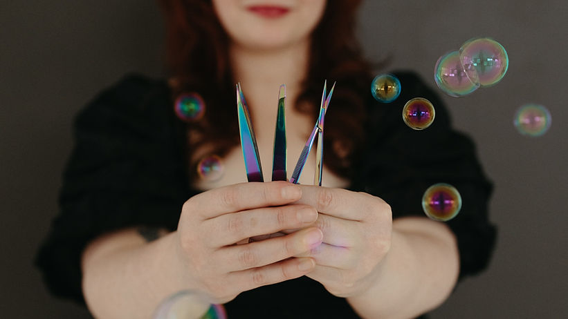 Closeup of esthetician hands holding two tweezers and bro w scissors wit bubbles in the foreground