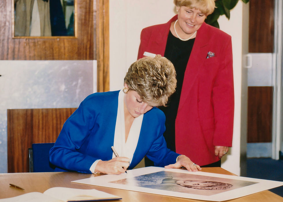 Princess Diana is seated at a table signing her portrait and Ruth Sims stands beside her.