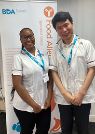 Two Mildmay student dietitians smiling in front of a BDA Food Allergy Specialist Group banner, wearing white clinical uniforms and event lanyards.