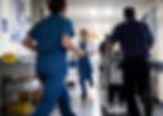 A busy NHS hospital ward with staff on the move and a porter wheeling an elderly patient in a wheelchair.