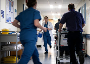 A busy NHS hospital ward with staff on the move and a porter wheeling an elderly patient in a wheelchair.