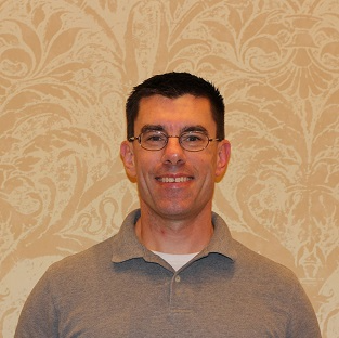 Smiling man wearing glasses and gray shirt against patterned background