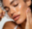 A close-up of a woman's face with glowing skin, showcasing freckles. She gently holds her face with one hand while applying a serum dropper near her cheek.