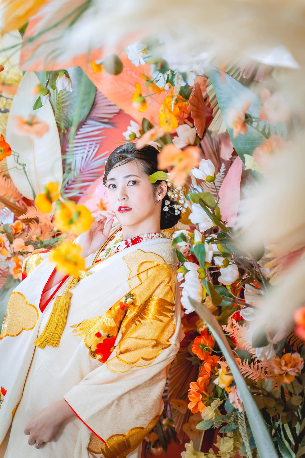 花に囲まれた白地に金模様の着物姿の女性が、正面を見つめる。華やかな花飾りの背景で、上品で幻想的な雰囲気。