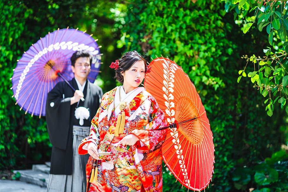 緑の庭園で、華やかな打掛の女性と黒い羽織の男性が和傘を手に並ぶ。赤と紫の傘が鮮やかで、静かな和装撮影の雰囲気。