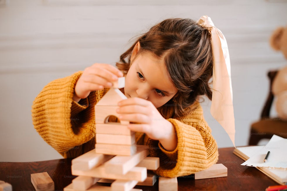 A child in a mustard sweater focuses on building a block tower at a table. Warm, cozy setting with soft lighting and a teddy bear in the background.