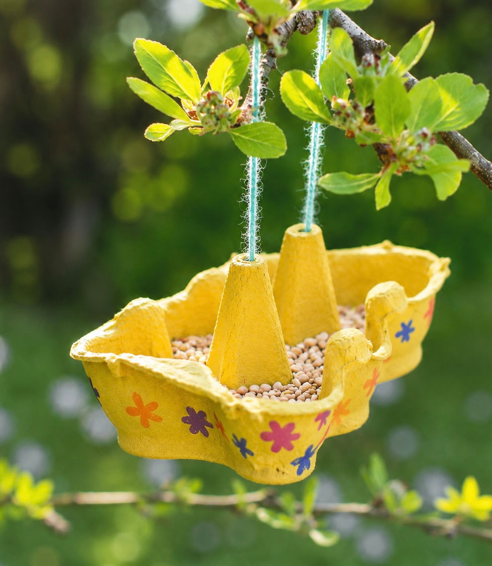Yellow homemade hanging bird feeder filled with seeds, suspended from a leafy tree branch against a blurred green garden.