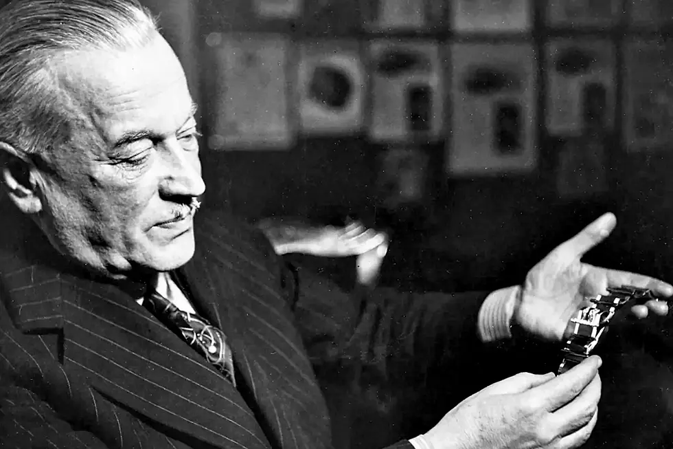 A black and white archive photo shows a watchmaker presenting a wristwatch inside a historic office setting.