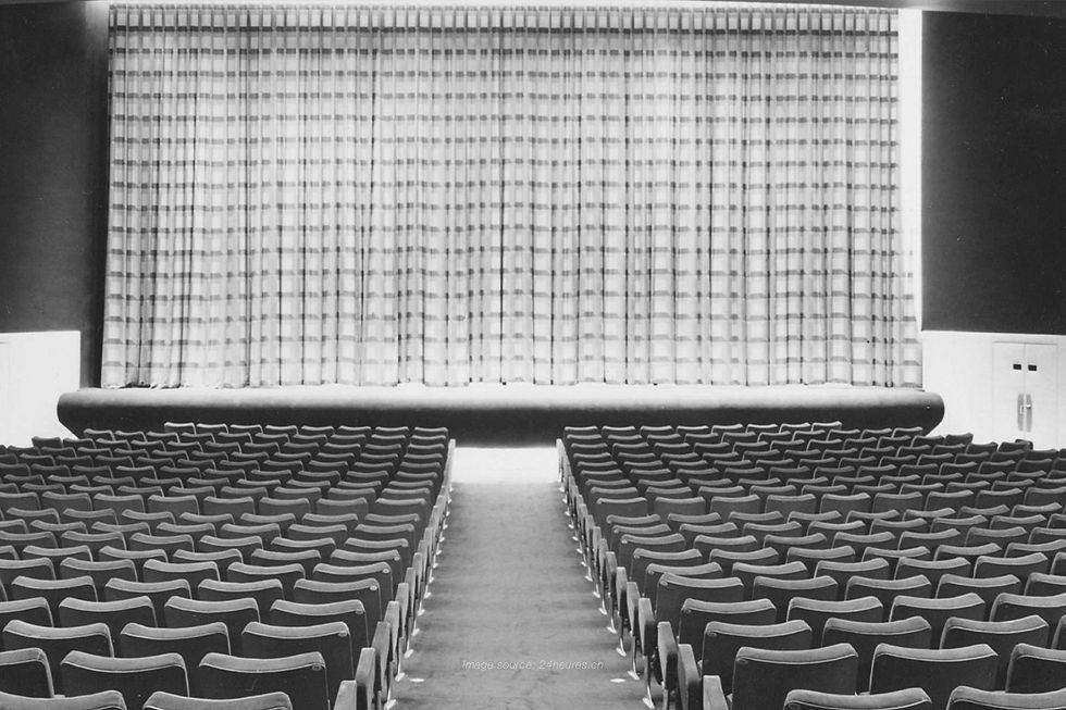Black and white photo of a classic cinema hall with rows of empty seats facing a large stage curtain.
