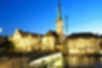 Fraumünster Church in Zurich illuminated at dusk beside historic buildings, a bridge, and the calm Limmat River.