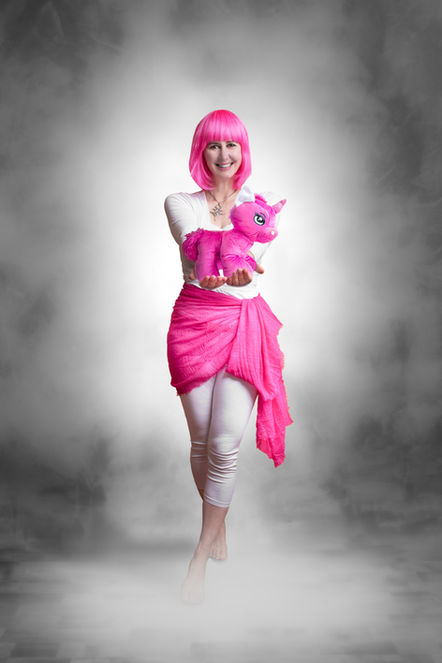 Une femme avec des cheveux rose et très souriante ,marche vers nous en tenant dans ses mains une licorne rose en peluche comme pour nous l'offrir. Le femme semble venir d'un autre monde à travers de la fumée, et semble flotter sur un nuage