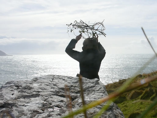 MFA artist and Clinical Herbalist Donna Troy Cleary performing a healing ceremony on the cliff edge at Cill Rialaig artist residency, County Kerry Ireland — holding a hand-woven bramble basket above her head against a silver Atlantic Ocean, conjuring Irish ancestors lost in the Famine, April 2017