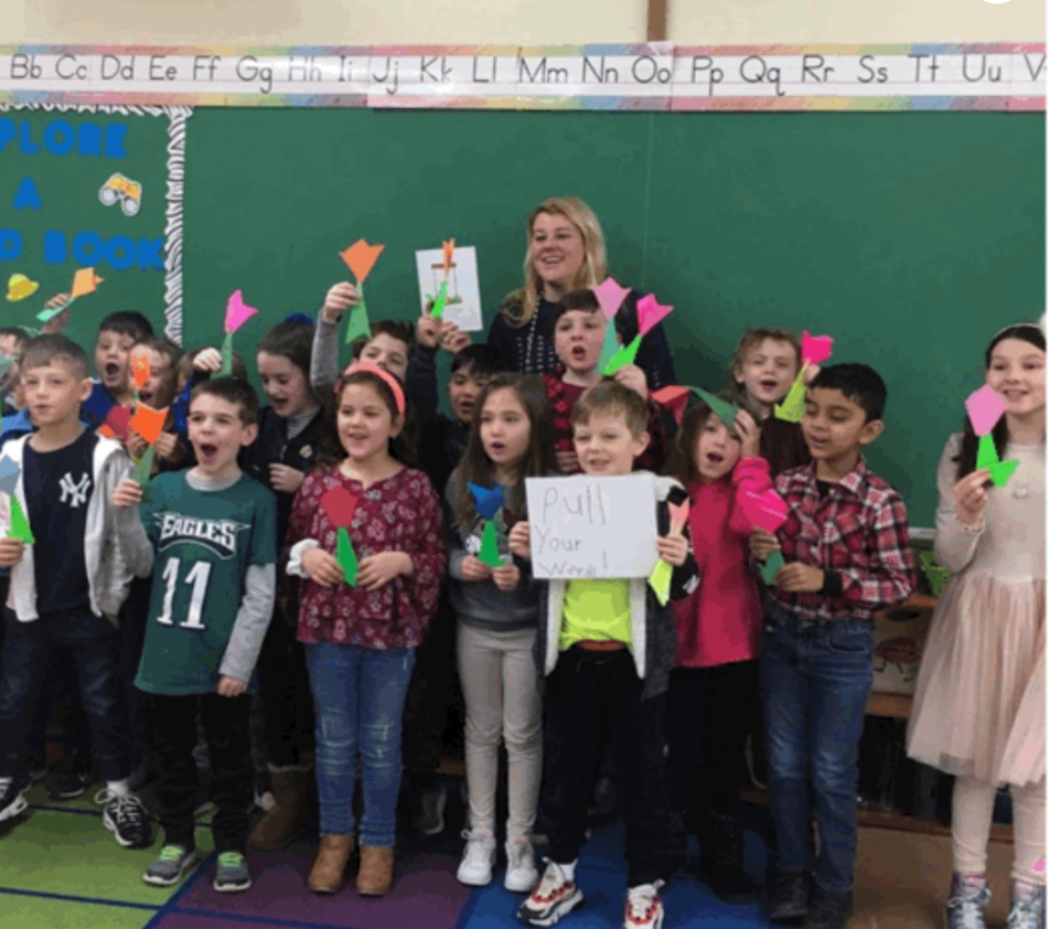 Gardener Shanna and smiling children in a classroom hold colorful paper shapes and signs, posing in front of a green board.