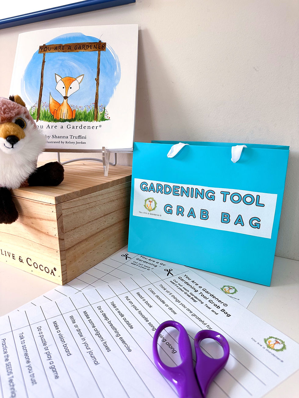 Blue bag labeled "Gardening Tool Grab Bag," book with a fox illustration, plush toy, and activity sheets on a table with purple scissors.