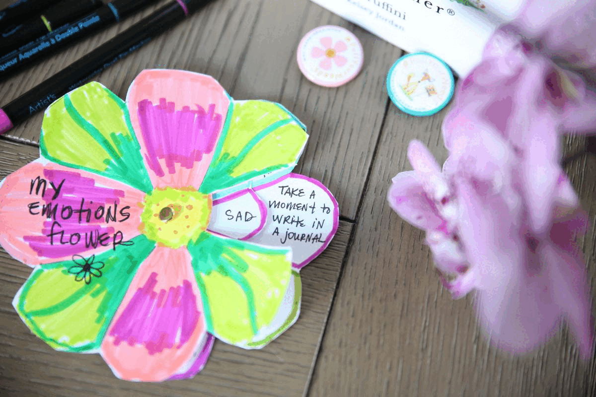 Colorful paper flower labeled "my emotions flower," with words "sad" and "take a moment to write in a journal." Markers and purple flowers on wood.