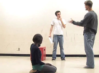 Scott Wallin speaks to a student, who listens, holding a script in one hand and brings his finger up to his chin. Another student kneels on the floor in the forground.
