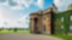 Sibton Park Manor, a grand mansion with ivy-covered walls, tall columns, and arched entrance. Surrounded by lush green grass and open skies with fluffy clouds.