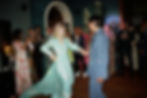 A couple dances in a vibrant room. The woman wears a light blue dress; the man is in a blue suit. Guests watch and take photos, smiling.