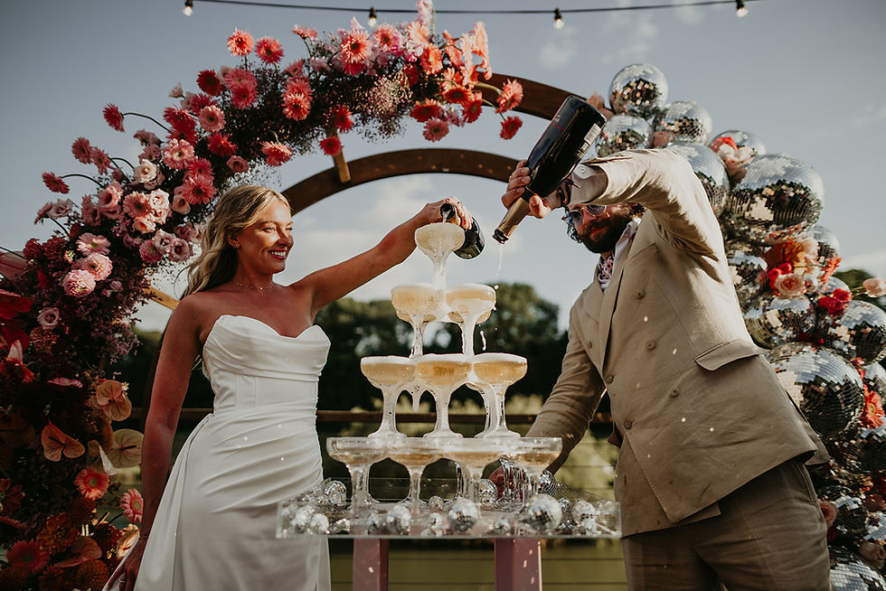 Bride and groom pours champagne into a tower of glasses under a floral and disco ball arch outdoors, with a festive, joyful mood.
