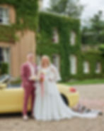 A couple in wedding attire stands smiling by a yellow convertible. Ivy-covered house, confetti, and attached cans in the background. Romantic mood.