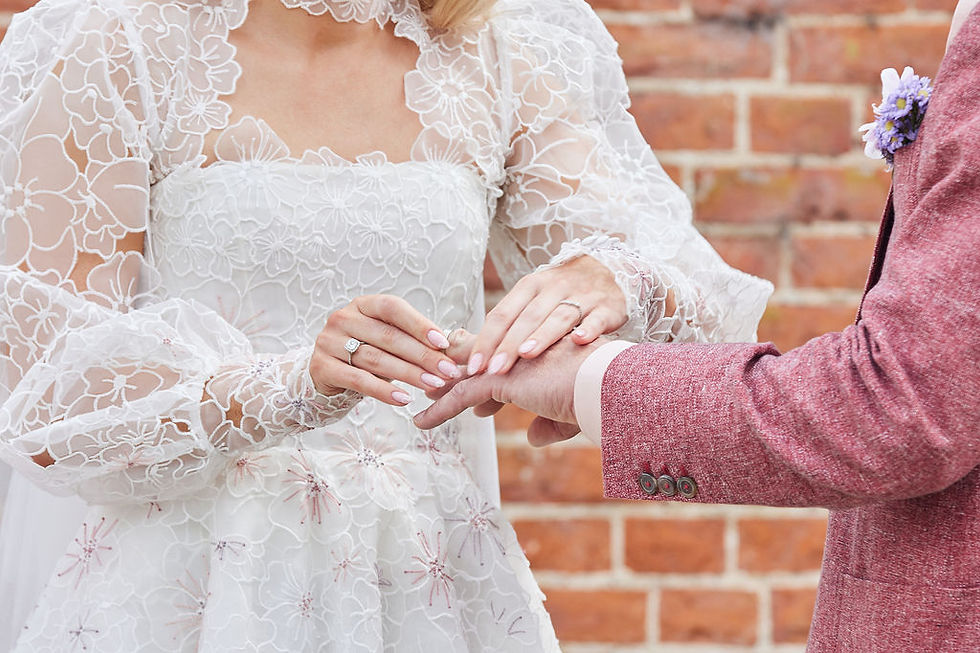 Bride in floral lace gown places a ring on groom's finger. Groom wears a pink jacket with a flower. Brick wall background. Romantic mood.
