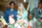 Smiling wedding couple at a floral-adorned table, woman in lace veil holding a glass, wood-panelled background. Festive, cheerful atmosphere.