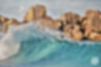 A turquoise wave crashes against a shoreline with rugged, layered rock formations in the background.